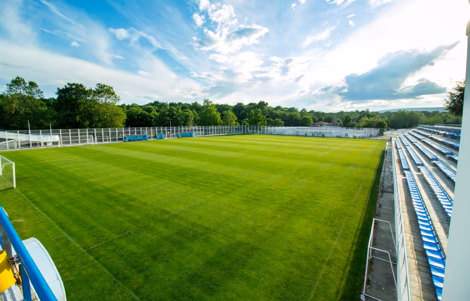 Empty soccer stadium with bright green grass field, blue seating stands, and a partly cloudy sky.